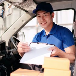 A man in a blue shirt is smiling and writing on a clipboard. He is driving a vehicle and he is a delivery driver